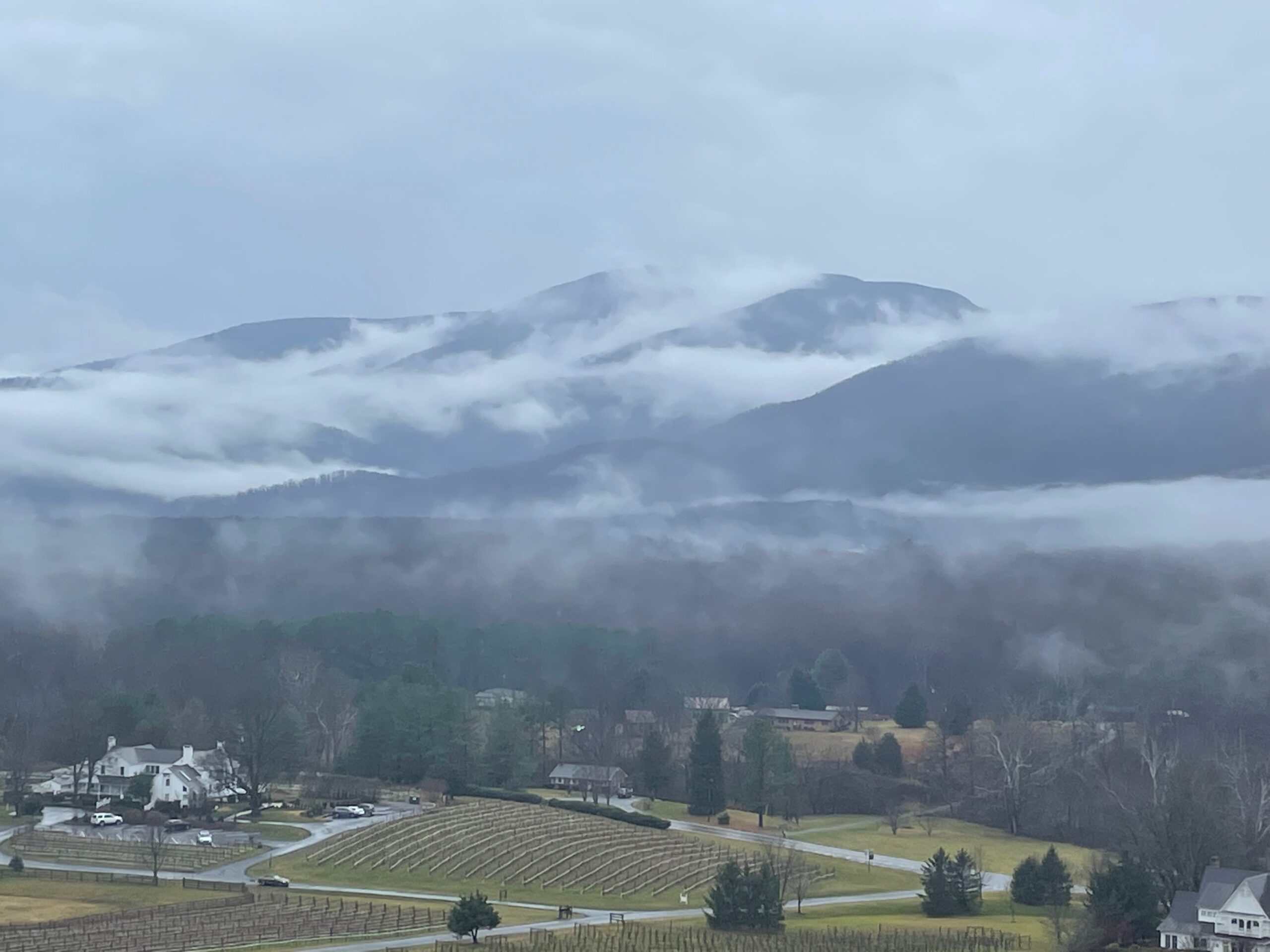 Winter landscape of the farmhouse at Veritas from a high viewpoint. Clouds cover mountains in the background.