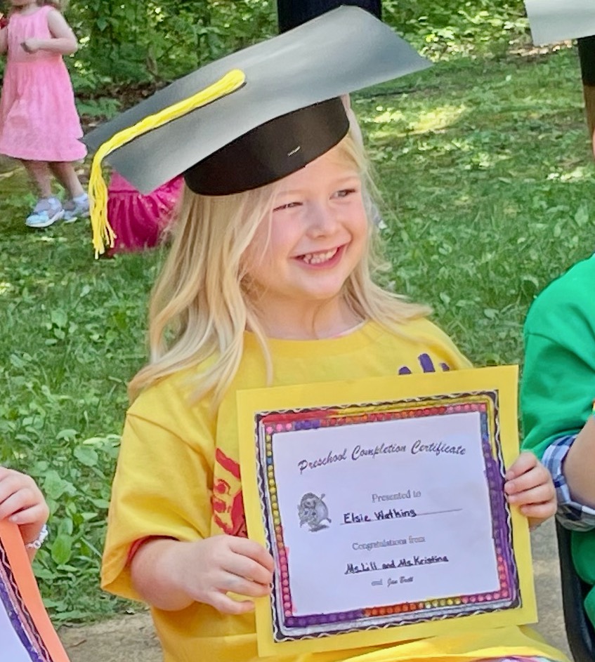 Photo of young girl in a paper graduation cap, wearing a yellow shirt, holding a Preschool Completion Certificate. 