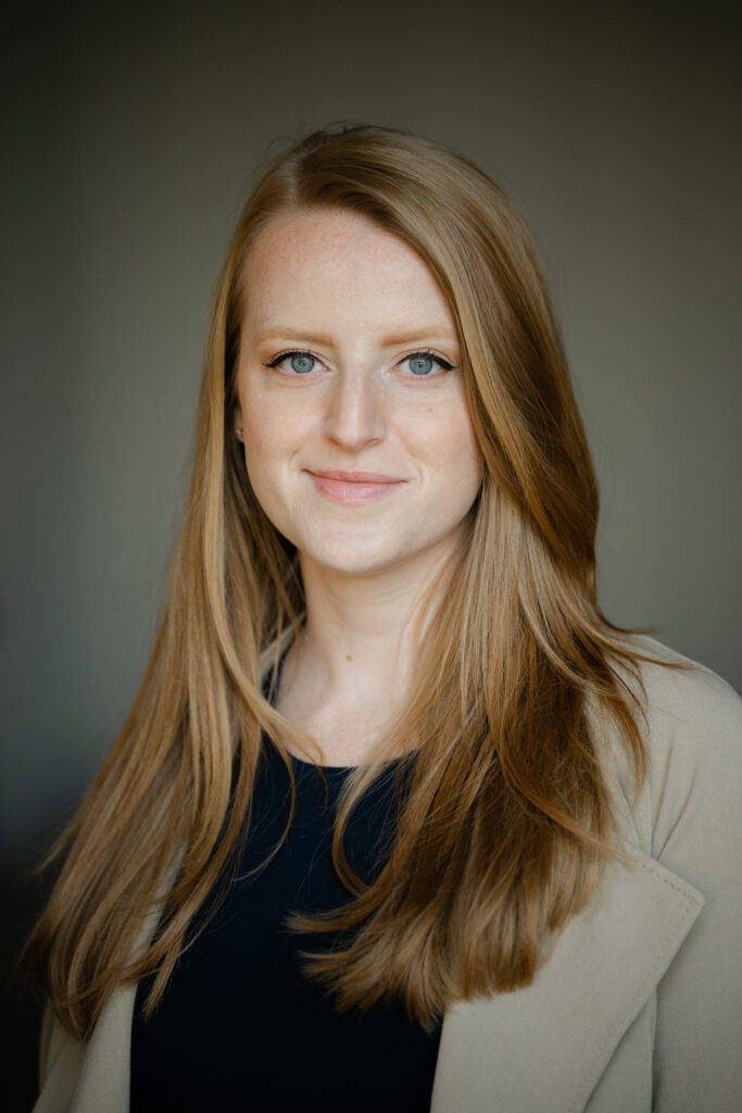 Headshot of a redheaded woman in a navy dress and tan jacket. 