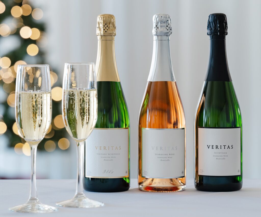 Photo of the Veritas Sparkling wine line up, including two flutes of sparkling wine, a bottle of Veritas Vintage Scintilla  2016, a bottle of Veritas Sparkling Rose, and a bottle of Veritas Scintilla non-vintage. 