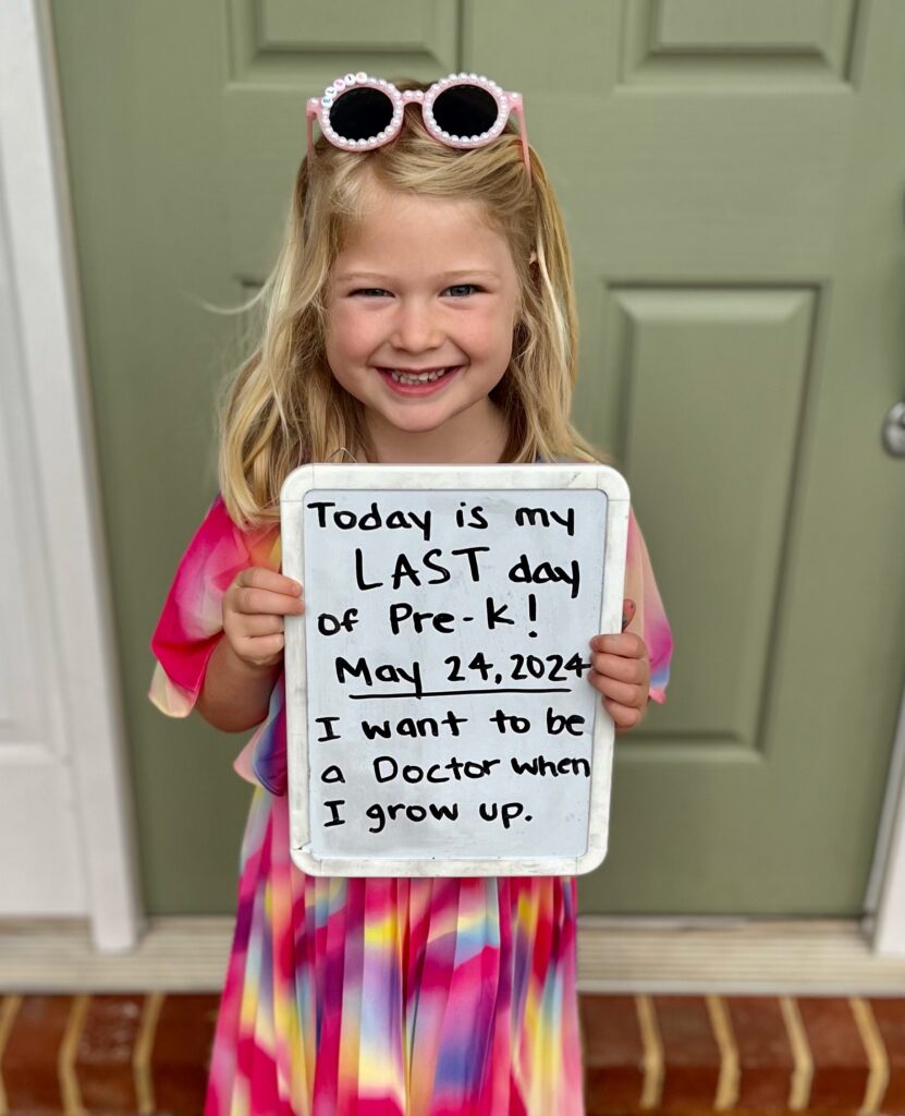 Photo of young girl with a colorful dress and sunglasses on her head holding a sign that says "Today is my LAST day of Pre-K! May 24, 2024. I want to be a doctor when I grow up."