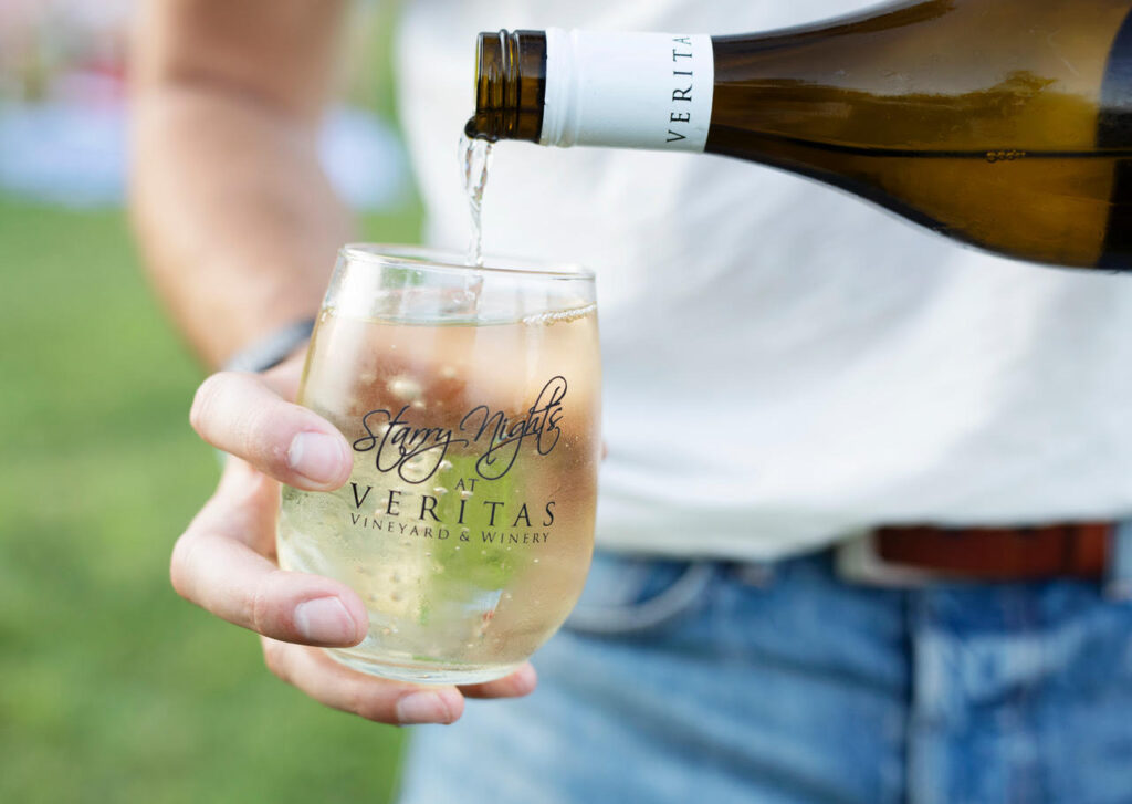 Photo of a person holding a stemless glass of wine as they fill it with white wine. 