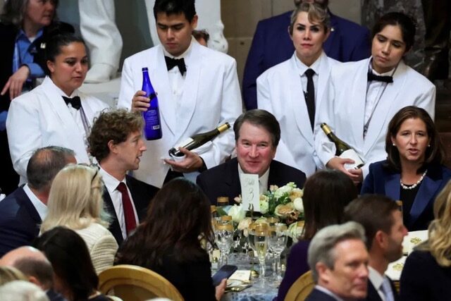 a group of waiters stand behind a table of guests, serving bottles of water and wine. 