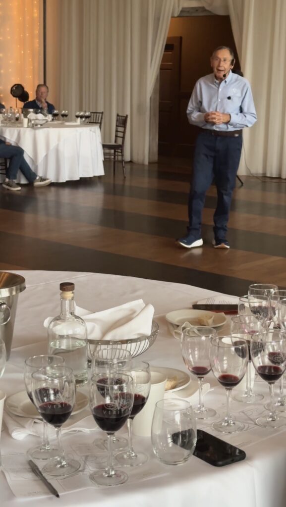 A man leads a group of people through a wine tasting in a large ballroom. Glasses of red wine cover tables. 