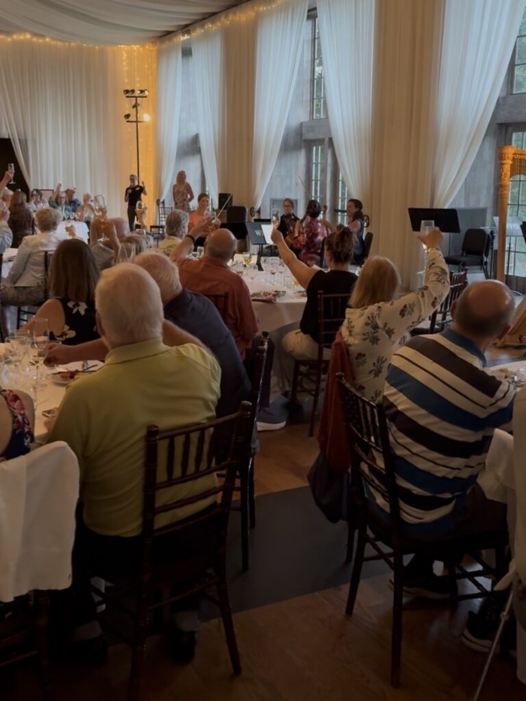 A large group of people sit in a ballroom, toasting to a group of musicians. 